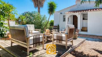 Mobilier de terrasse en bois avec coussins blancs sur une terrasse en bois devant la Villa Sofia, caractérisée par un mur extérieur blanc, des tuiles de terre cuite et des palmier sous un ciel bleu dégagé.