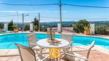 Espace repas en plein air près de la piscine de la Villa Sofia, doté d'une table blanche en plastique, de quatre chaises assorties avec coussins et d'un centre de table avec une lanterne en osier, offrant une vue sur les collines lointaines.