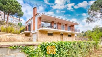Vue extérieure de la Villa Lloret mettant en valeur une maison à deux étages avec un rez-de-chaussée en pierre, un étage supérieur en brique et un balcon filant sous un ciel bleu nuageux.