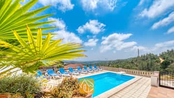 Piscine privée avec des transats bleus et une palme de cocotier au premier plan à la Villa Santa Maria, sous un ciel bleu parsemé de quelques nuages.