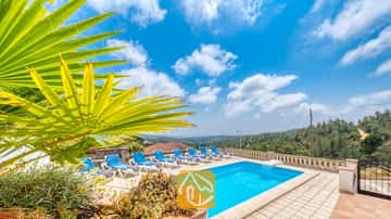 Piscine privée avec des transats bleus et une palme de cocotier au premier plan à la Villa Santa Maria, sous un ciel bleu parsemé de quelques nuages.