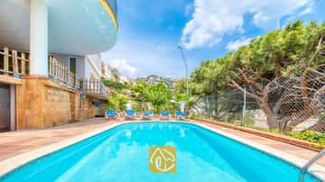 Piscine extérieure de la Villa Valentina aux eaux bleues, entourée d'un sol carrelé, de cinq chaises longues et de pins sous un ciel bleu.