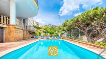 Piscine extérieure de la Villa Valentina aux eaux bleues, entourée d'un sol carrelé, de cinq chaises longues et de pins sous un ciel bleu.