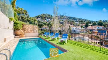 Piscine privée avec des transats bleus sur une terrasse en gazon synthétique verdoyant à la Villa Donna, surplombant les maisons à flanc de colline sous un ciel d'un bleu éclatant.