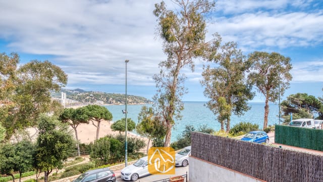 Vue depuis Casa Costa montrant de grands arbres, une plage de sable et la mer bleue sous un ciel partiellement nuageux.