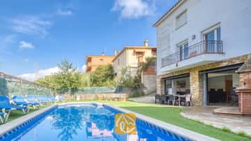 Piscine privée et terrasse de la Villa Nicky, dotées de bains de soleil bleus sur gazon synthétique, le long d'une villa blanche à deux étages agrémentée de colonnes en pierre et de baies vitrées coulissantes.