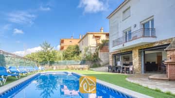 Piscine privée et terrasse de la Villa Nicky, dotées de bains de soleil bleus sur gazon synthétique, le long d'une villa blanche à deux étages agrémentée de colonnes en pierre et de baies vitrées coulissantes.