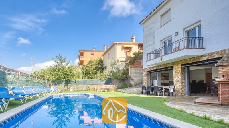 Piscine privée et terrasse de la Villa Nicky, dotées de bains de soleil bleus sur gazon synthétique, le long d'une villa blanche à deux étages agrémentée de colonnes en pierre et de baies vitrées coulissantes.