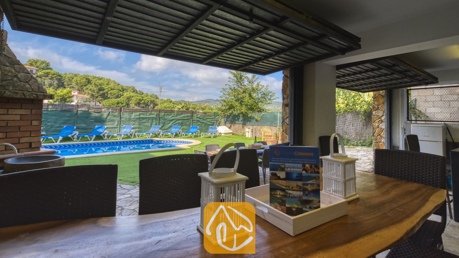 Espace repas extérieur couvert à la Villa Nicky, doté d'une table en bois agrémentée de lanternes blanches, offrant une vue sur la piscine et la pelouse verdoyante équipée de chaises longues.
