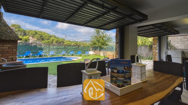 Espace repas extérieur couvert à la Villa Nicky, doté d'une table en bois agrémentée de lanternes blanches, offrant une vue sur la piscine et la pelouse verdoyante équipée de chaises longues.