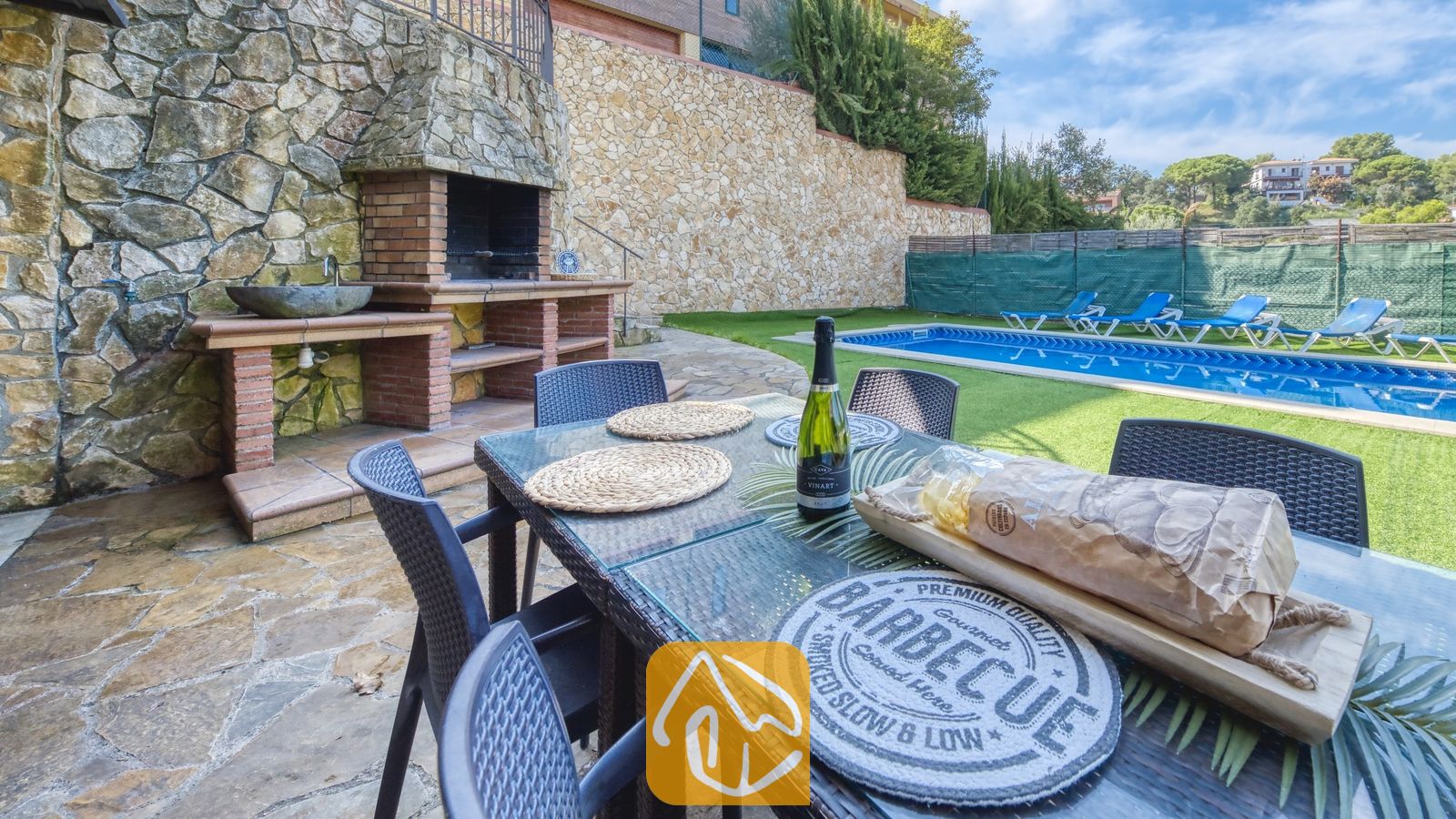 Table à manger extérieure dressée avec du pain et une bouteille de vin à la Villa Nicky, dotée d'un barbecue en pierre intégré et d'une piscine en arrière-plan.