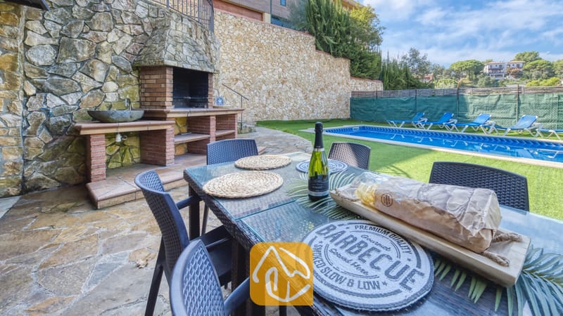 Table à manger extérieure dressée avec du pain et une bouteille de vin à la Villa Nicky, dotée d'un barbecue en pierre intégré et d'une piscine en arrière-plan.