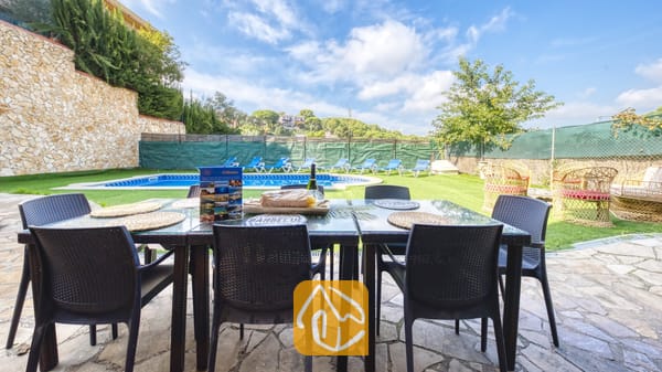 Table à manger extérieure installée sur une terrasse en pierre à la Villa Nicky, surplombant une piscine, une pelouse et un ciel bleu parsemé de nuages blancs.