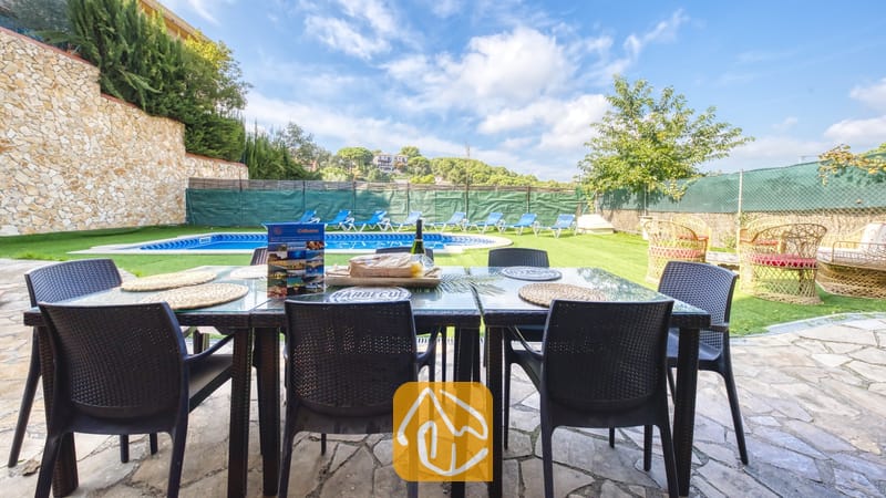 Table à manger extérieure installée sur une terrasse en pierre à la Villa Nicky, surplombant une piscine, une pelouse et un ciel bleu parsemé de nuages blancs.