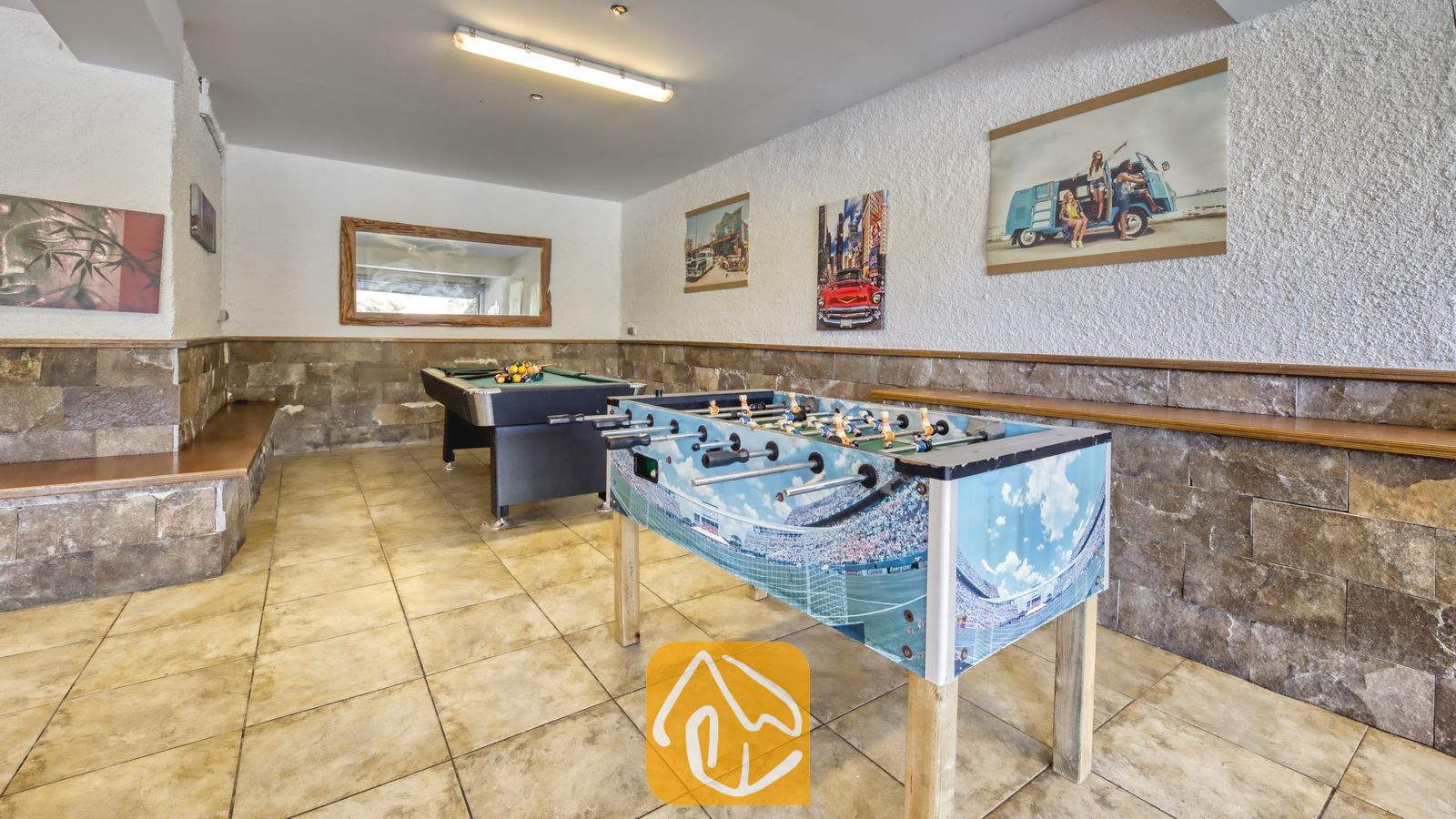 Salle de jeux de la Villa Nicky comprenant, au premier plan, un baby-foot avec des graphismes de stade, un billard en arrière-plan, des murs en pierre et des œuvres d'art encadrées.