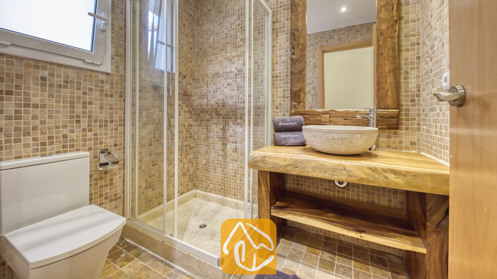 Salle de bains dotée d'une douche d'angle vitrée, d'une vasque en pierre posée sur un meuble sous-lavabo en bois brut aux bords naturels, et de murs en mosaïque beige.