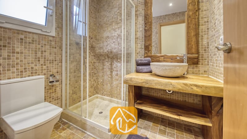 Salle de bains dotée d'une douche d'angle vitrée, d'une vasque en pierre posée sur un meuble sous-lavabo en bois brut aux bords naturels, et de murs en mosaïque beige.