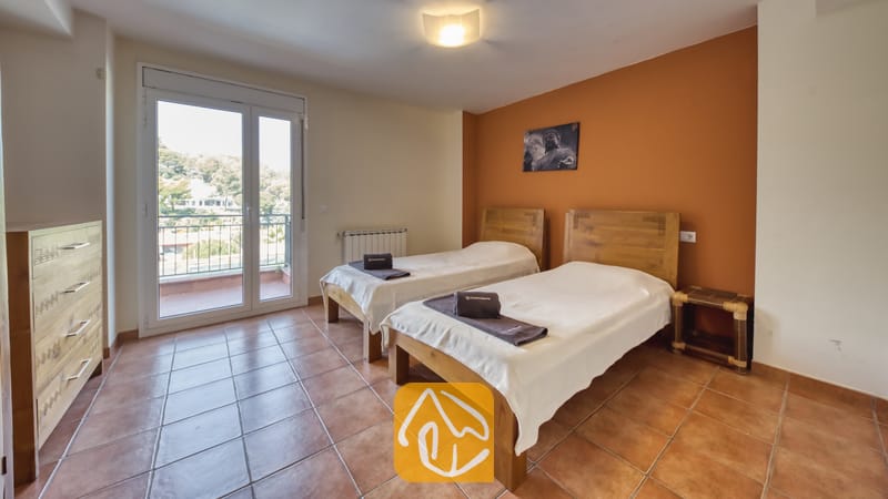 Chambre à lits jumeaux dotée de têtes de lit en bois, de serviettes sombres roulées sur des dessus-de-lit blancs, de sols carrelés et d'une porte-fenêtre ouvrant sur des arbres.