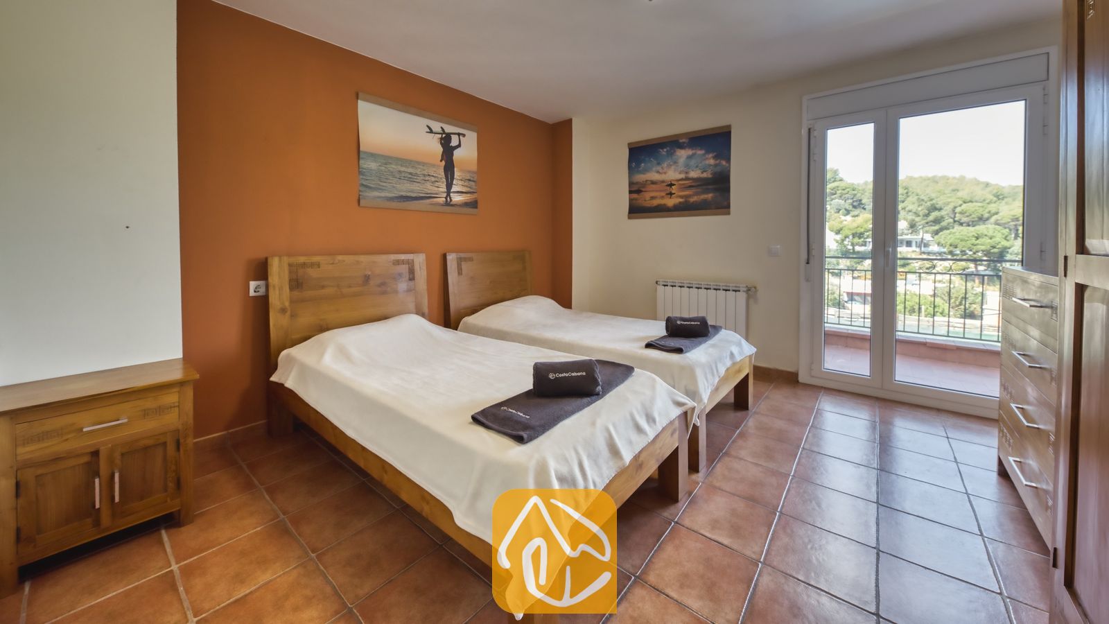 Chambre à lits jumeaux de la Villa Nicky comprenant deux lits en bois, du linge de lit blanc, des sols en terre cuite et une porte-fenêtre donnant sur des collines verdoyantes.