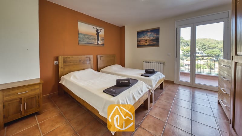 Chambre à lits jumeaux de la Villa Nicky comprenant deux lits en bois, du linge de lit blanc, des sols en terre cuite et une porte-fenêtre donnant sur des collines verdoyantes.