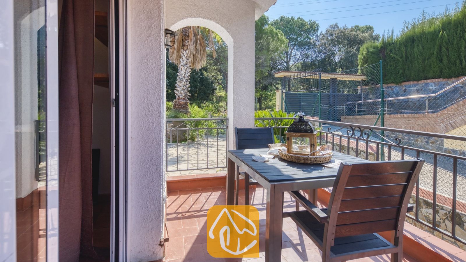 Balcon de la Villa Nicky doté d'une table à manger en bois sombre, de chaises, d'une lanterne décorative et d'un sol carrelé, offrant une vue sur les arbres et les clôtures.