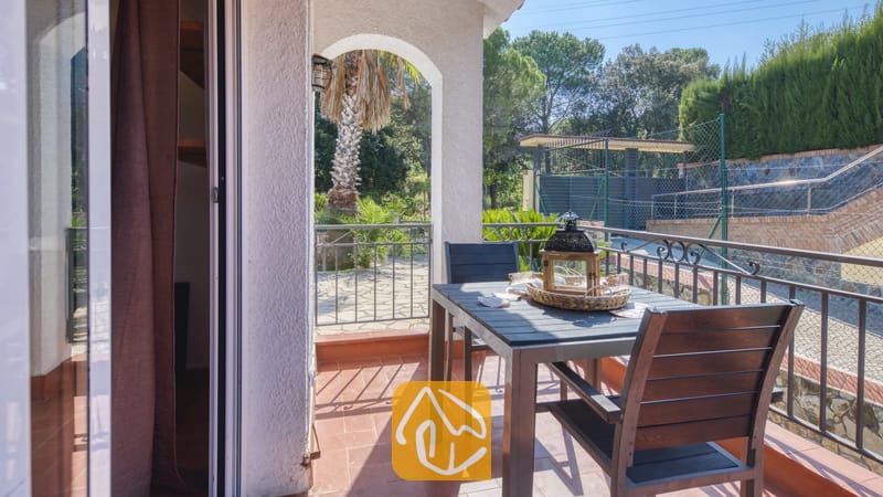 Balcon de la Villa Nicky doté d'une table à manger en bois sombre, de chaises, d'une lanterne décorative et d'un sol carrelé, offrant une vue sur les arbres et les clôtures.