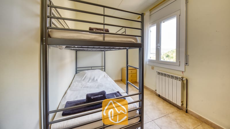 Chambre avec des lits superposés en métal gris, du linge de lit blanc, des serviettes foncées pliées, une petite table de chevet en bois et une fenêtre offrant une vue sur l'extérieur.