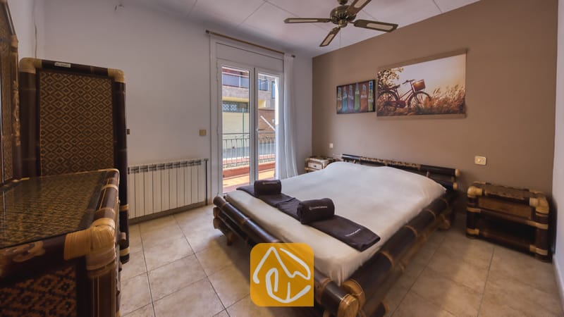 Chambre dotée d'un lit avec un cadre en bambou et des serviettes marron roulées, de sols carrelés, de mobilier assorti en bois foncé et de portes vitrées s'ouvrant sur une terrasse.