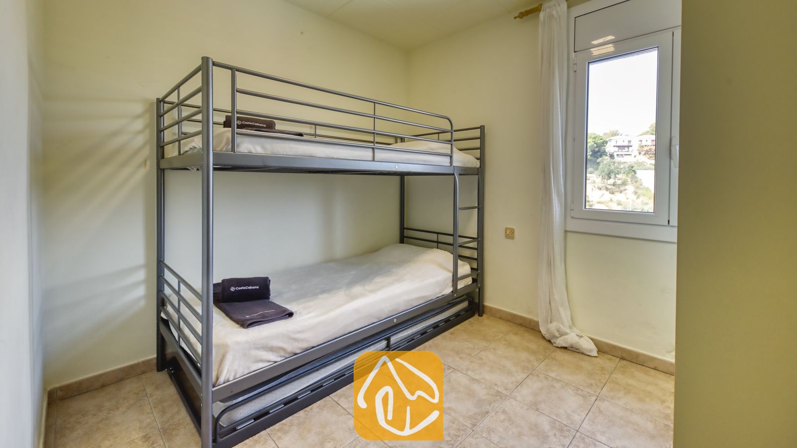 Chambre comprenant un lit superposé en métal gris avec du linge de lit blanc et des serviettes foncées pliées, un sol carrelé et une petite fenêtre avec des rideaux blancs.