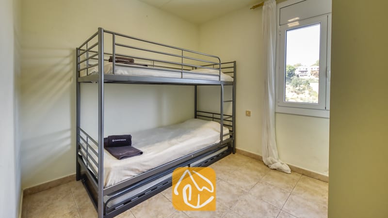 Chambre comprenant un lit superposé en métal gris avec du linge de lit blanc et des serviettes foncées pliées, un sol carrelé et une petite fenêtre avec des rideaux blancs.