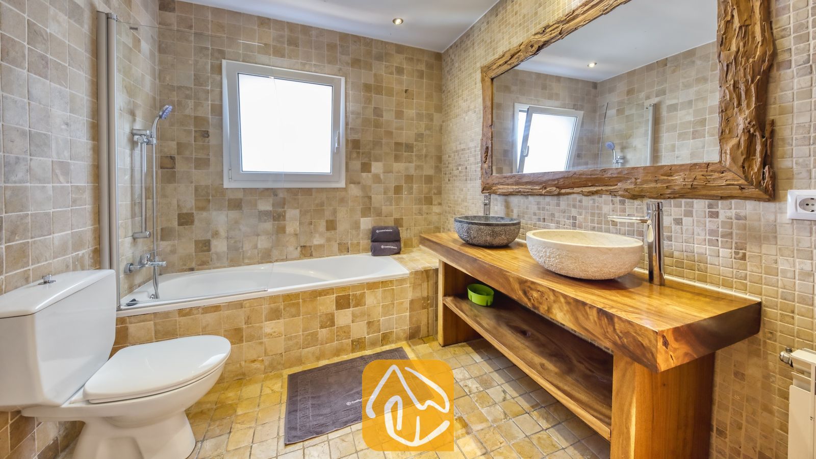 Salle de bains aux murs carrelés de beige, dotée d'une baignoire avec pommeau de douche, d'un meuble sous-vasque en bois surmonté de deux vasques en pierre, ainsi que d'un grand miroir pourvu d'un cadre en bois rustique.