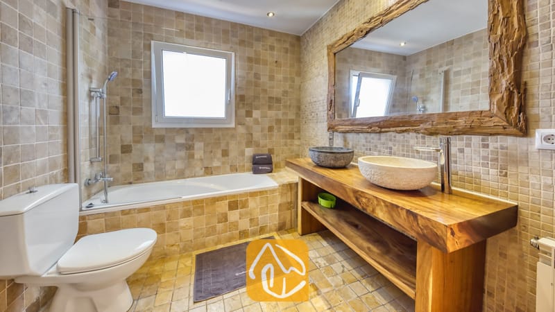 Salle de bains aux murs carrelés de beige, dotée d'une baignoire avec pommeau de douche, d'un meuble sous-vasque en bois surmonté de deux vasques en pierre, ainsi que d'un grand miroir pourvu d'un cadre en bois rustique.