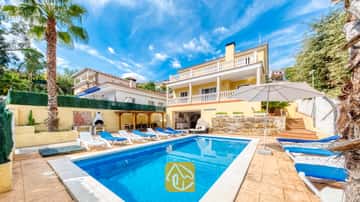 Piscine privée à la Villa Ashley, entourée de transats bleus et d'un parasol blanc, devant un bâtiment jaune de deux étages agrémenté de balustrades blanches.
