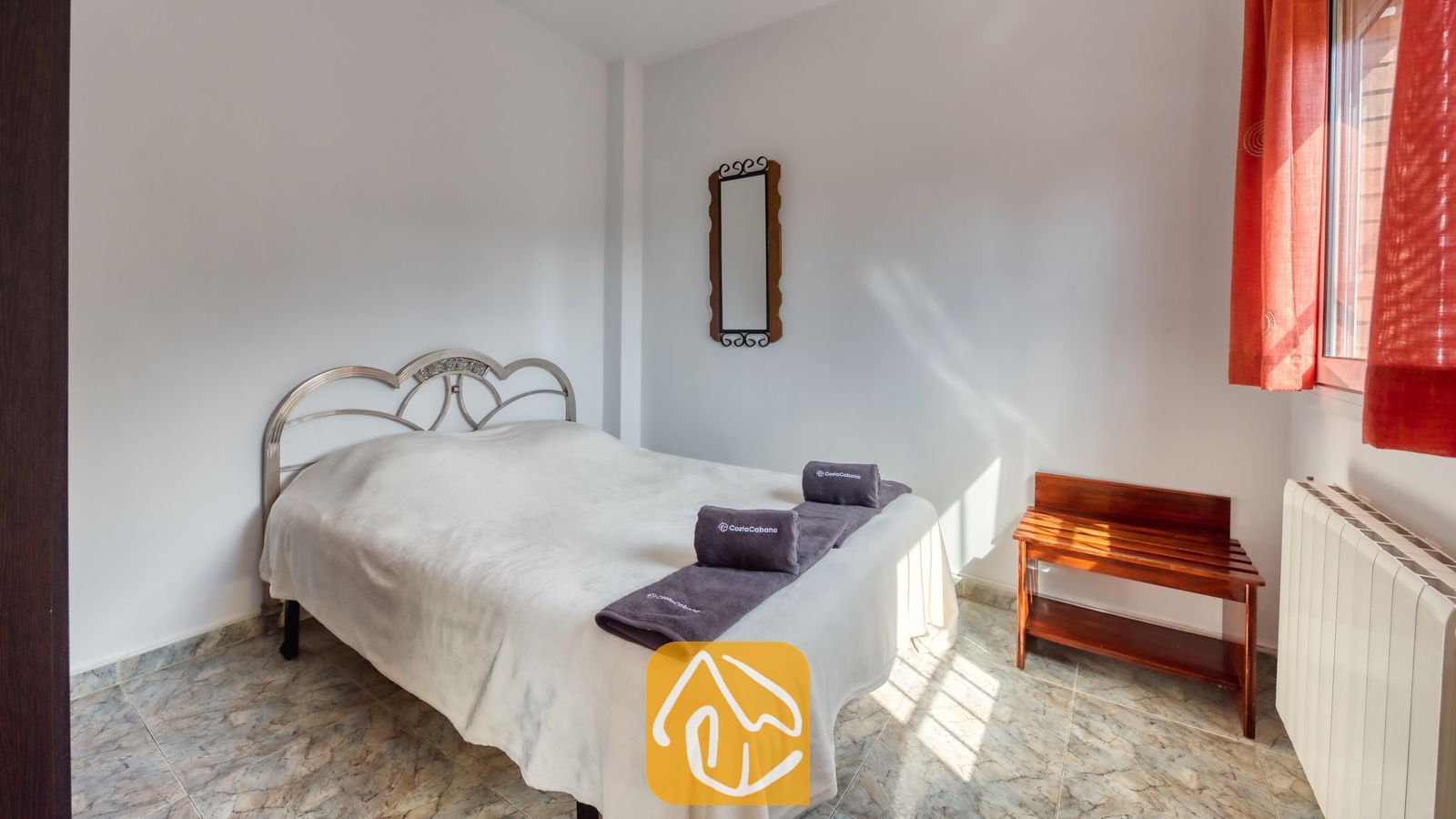 Chambre avec un lit blanc, des serviettes grises pliées, une tête de lit en métal, des rideaux rouges et un porte-bagages en bois.