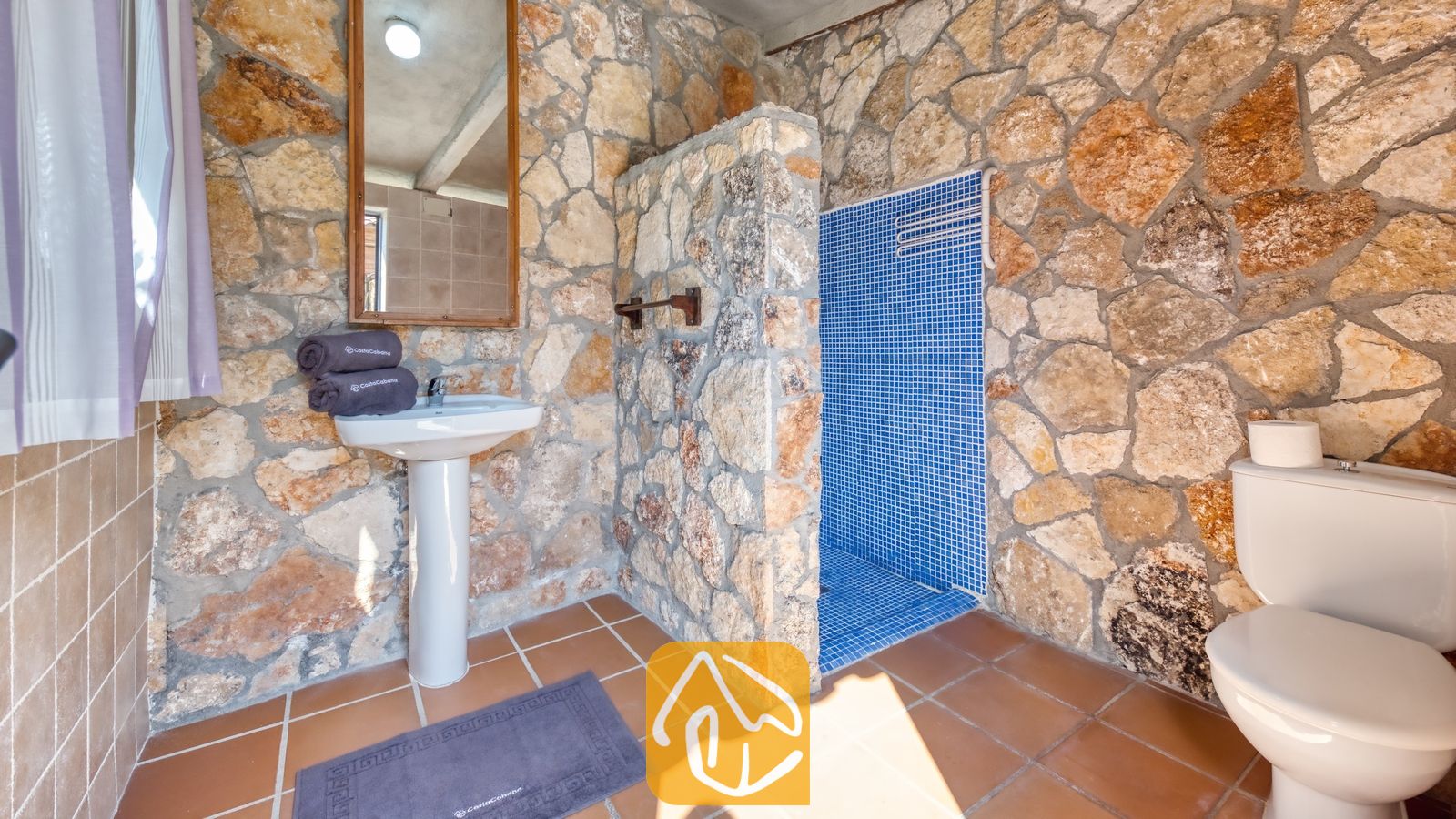 Salle de bains dotée de murs en pierre rustique, d'un lavabo sur pied blanc, d'une cabine de douche carrelée avec des mosaïques bleues et de carreaux de sol marron.