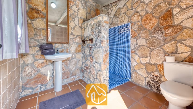 Salle de bains dotée de murs en pierre rustique, d'un lavabo sur pied blanc, d'une cabine de douche carrelée avec des mosaïques bleues et de carreaux de sol marron.