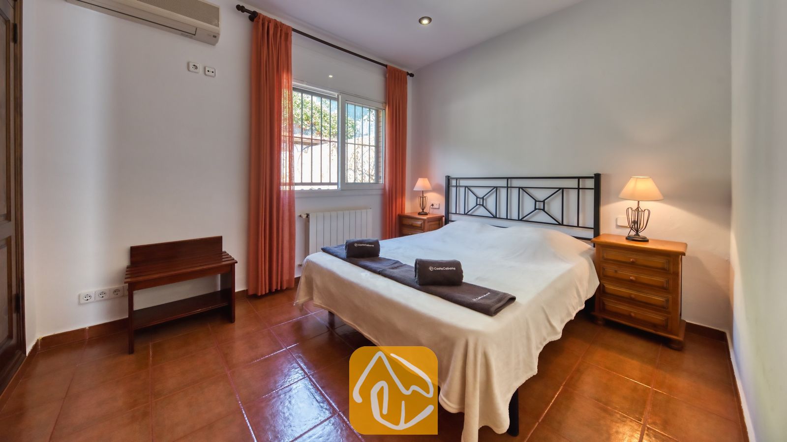 Chambre avec un lit double, un cadre de lit en métal foncé, des rideaux orange encadrant la fenêtre et deux tables de chevet en bois avec lampes.