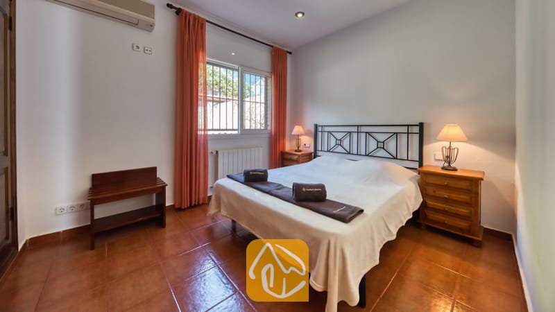 Chambre avec un lit double, un cadre de lit en métal foncé, des rideaux orange encadrant la fenêtre et deux tables de chevet en bois avec lampes.