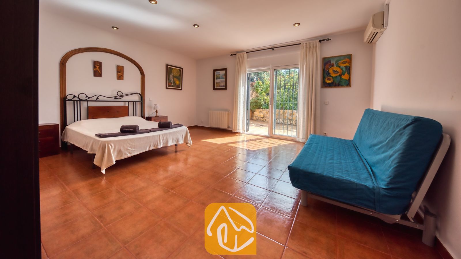 Spacieuse chambre à la Villa Alba dotée d'un lit double, d'un cadre de lit en métal foncé, d'un canapé bleu et de portes coulissantes en verre s'ouvrant sur une terrasse extérieure.