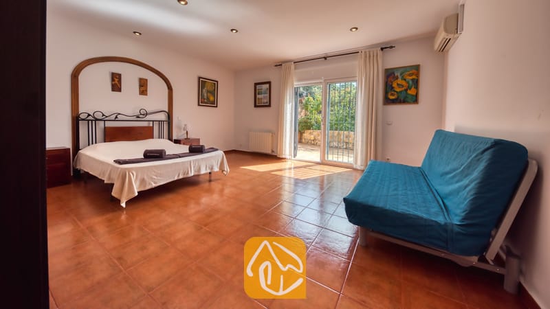 Spacieuse chambre à la Villa Alba dotée d'un lit double, d'un cadre de lit en métal foncé, d'un canapé bleu et de portes coulissantes en verre s'ouvrant sur une terrasse extérieure.