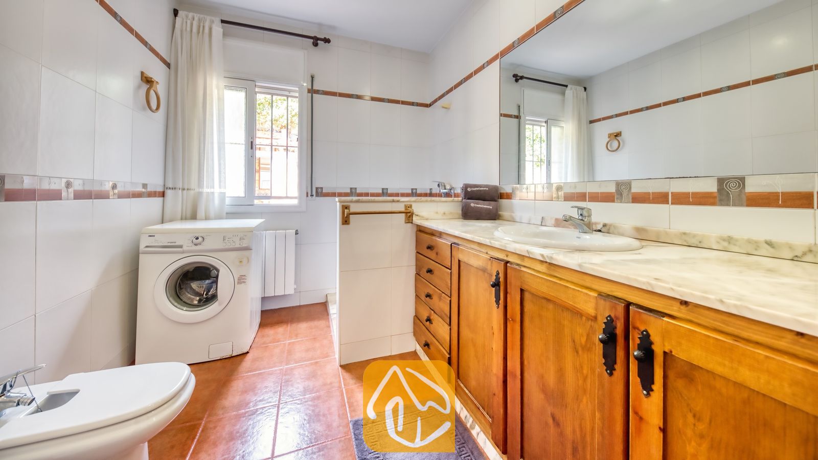 Salle de bains lumineuse et carrelée, dotée d'un meuble sous-vasque en bois avec plan de travail en marbre, d'un lave-linge blanc et de sols en terracotta.