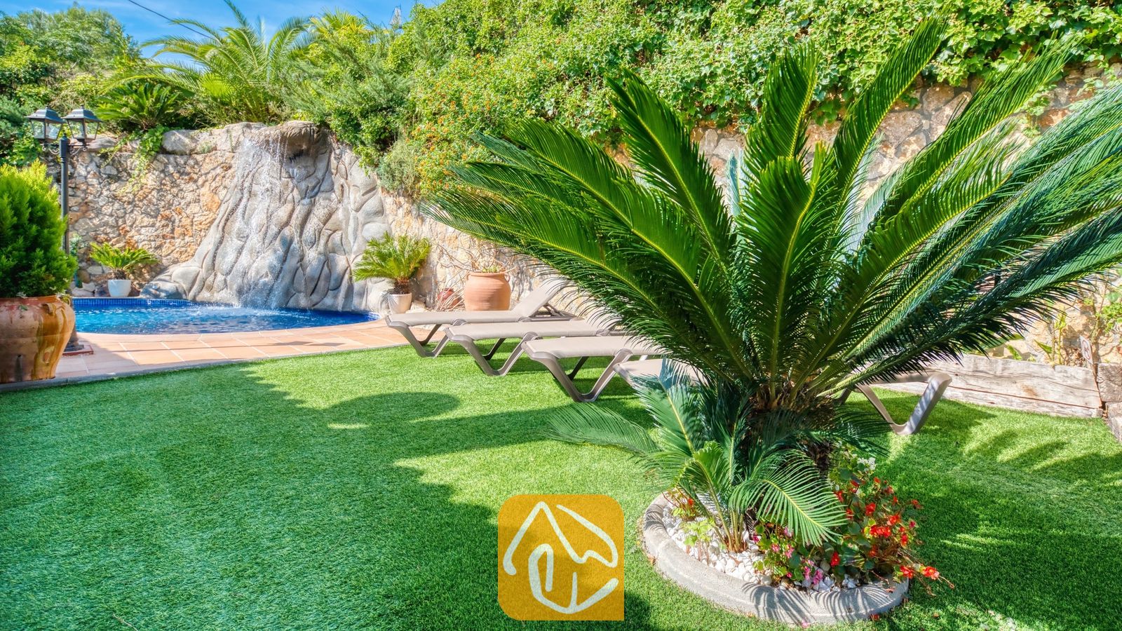 Jardin luxuriant et verdoyant de la Villa Alba, doté de gazon synthétique, d'un palmier cycas remarquable, de chaises longues et d'un plan d'eau en pierre agrémenté d'une cascade.