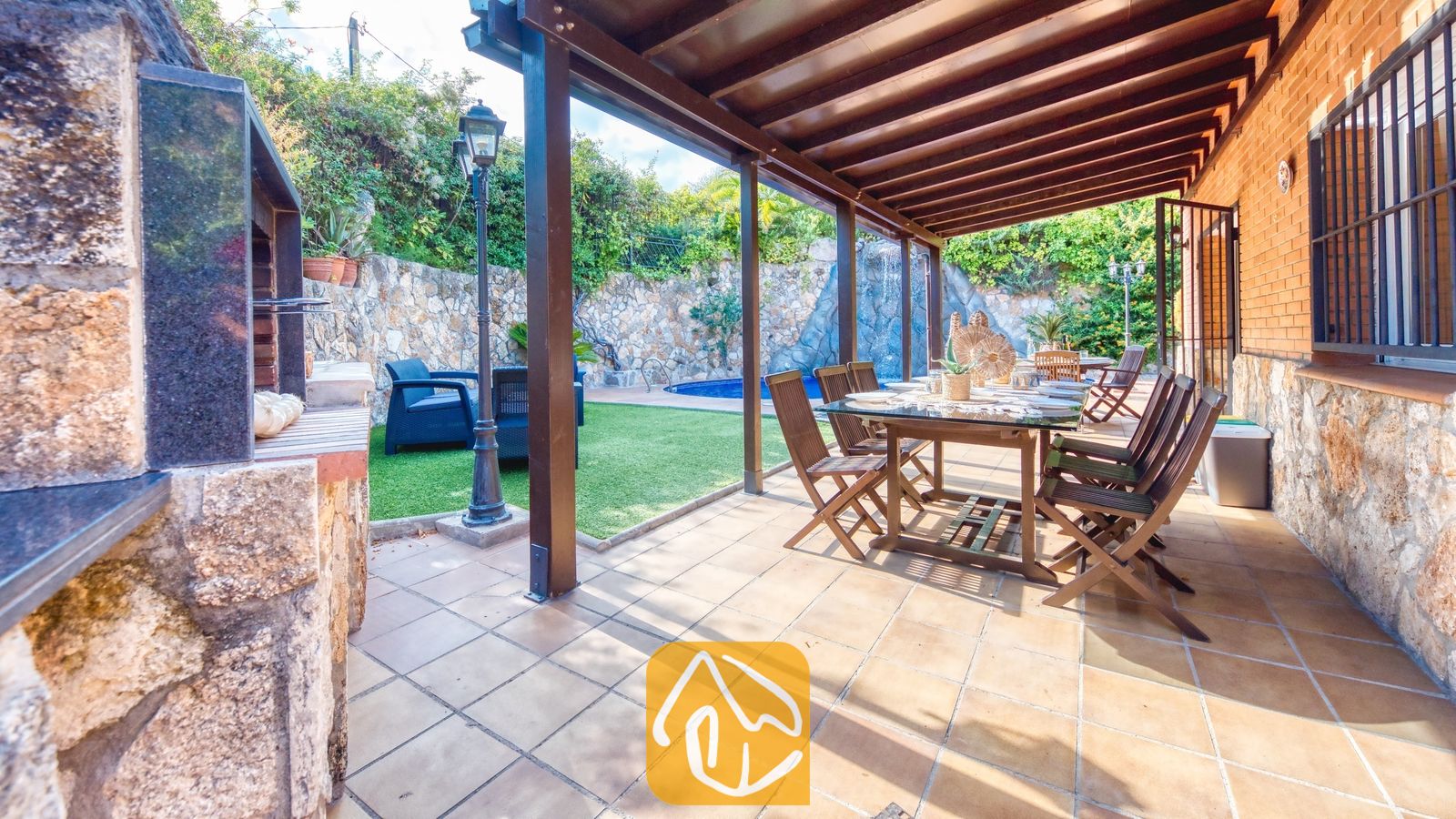 Terrasse repas extérieure couverte à la Villa Alba, dotée d'une table et de chaises de salle à manger en bois, d'un sol carrelé et d'un espace barbecue intégré attenant, offrant une vue sur le jardin et la piscine.
