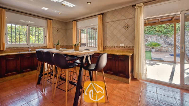 Coin repas de la cuisine doté d'une table haute en bois et de tabourets de bar noirs, de sols en terre cuite, de murs carrelés et d'une porte vitrée s'ouvrant sur une terrasse extérieure à la Villa Alba.