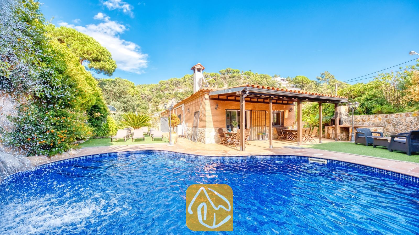 Piscine privée avec cascade en pierre à la Villa Alba, mettant en valeur une maison en briques dotée d'une terrasse couverte et de chaises longues sur pelouse artificielle sous un ciel bleu.