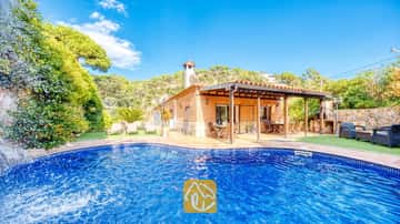 Piscine privée avec cascade en pierre à la Villa Alba, mettant en valeur une maison en briques dotée d'une terrasse couverte et de chaises longues sur pelouse artificielle sous un ciel bleu.