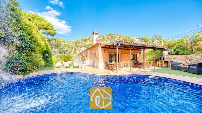 Piscine privée avec cascade en pierre à la Villa Alba, mettant en valeur une maison en briques dotée d'une terrasse couverte et de chaises longues sur pelouse artificielle sous un ciel bleu.