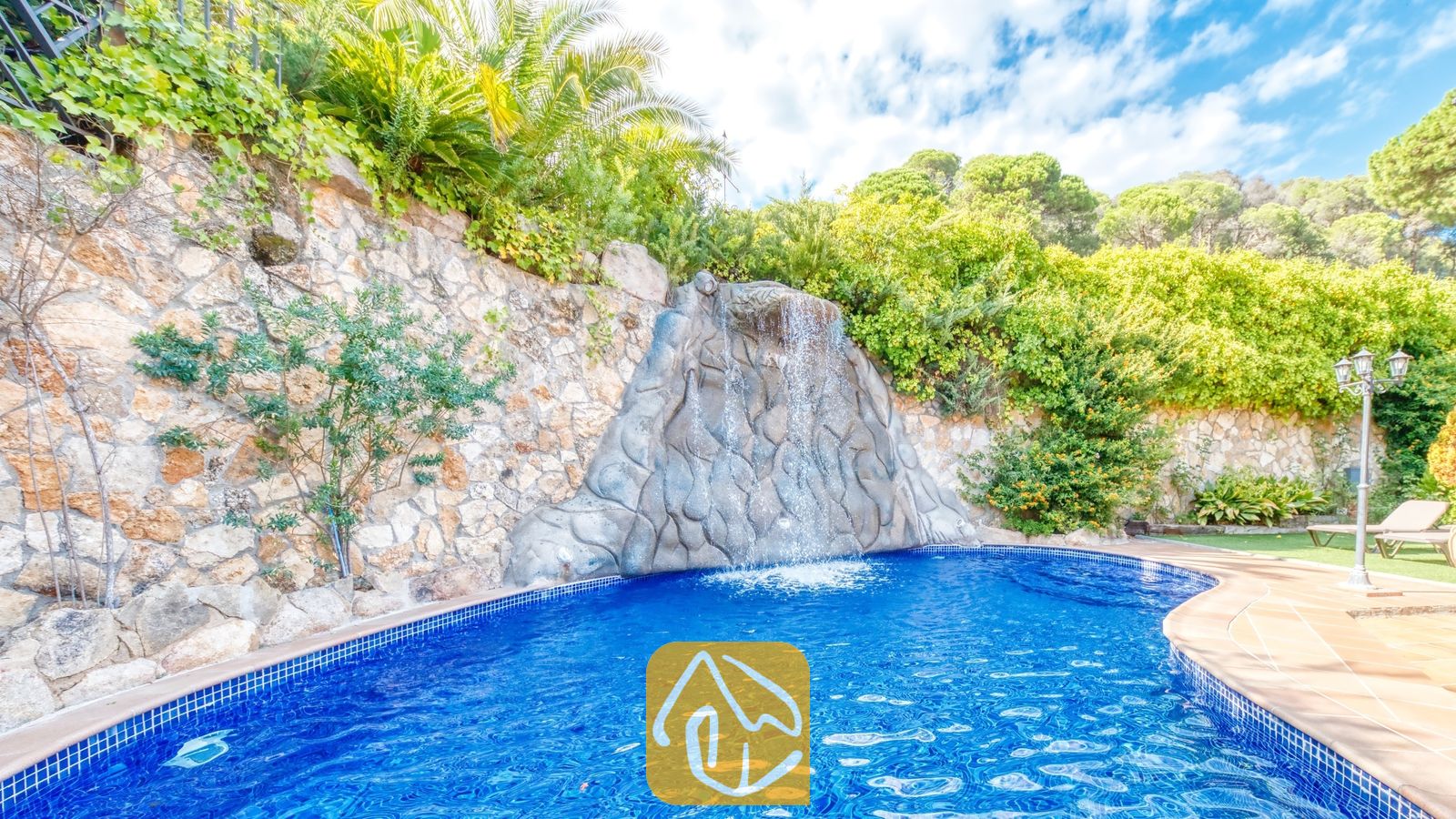 Cascade en rocaille se déversant dans une piscine carrelée de bleu à la Villa Alba, entourée de murs de soutènement en pierre et d'une végétation luxuriante sous un ciel partiellement nuageux.