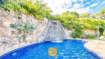 Cascade en rocaille se déversant dans une piscine carrelée de bleu à la Villa Alba, entourée de murs de soutènement en pierre et d'une végétation luxuriante sous un ciel partiellement nuageux.