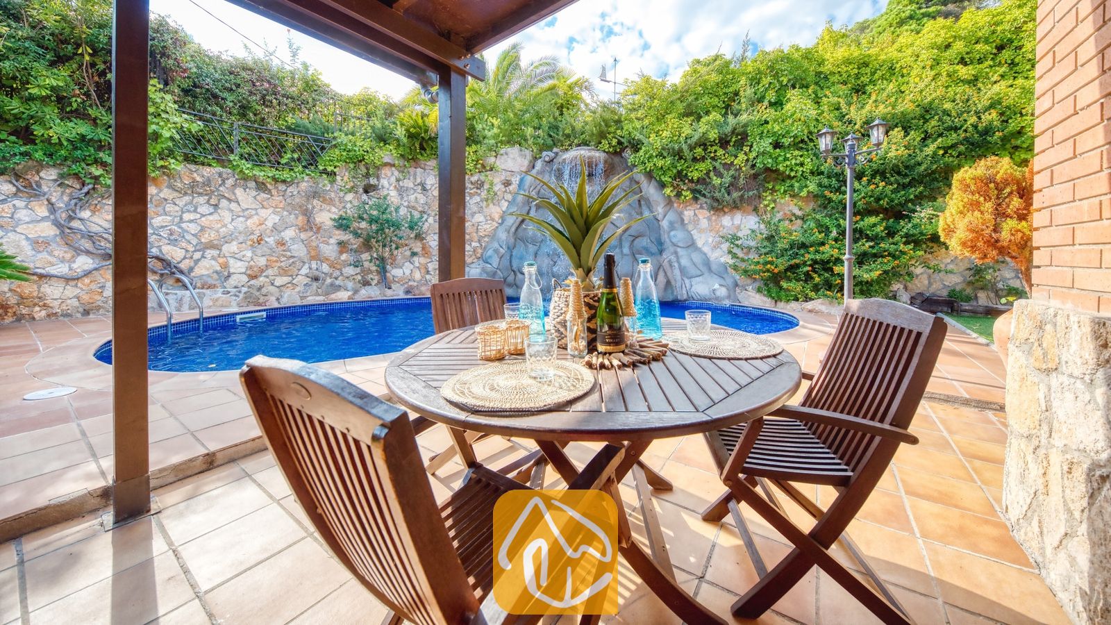 Table et chaises de repas en bois sur une terrasse carrelée à la Villa Alba, avec une piscine bleue et une cascade de rocailles en arrière-plan.